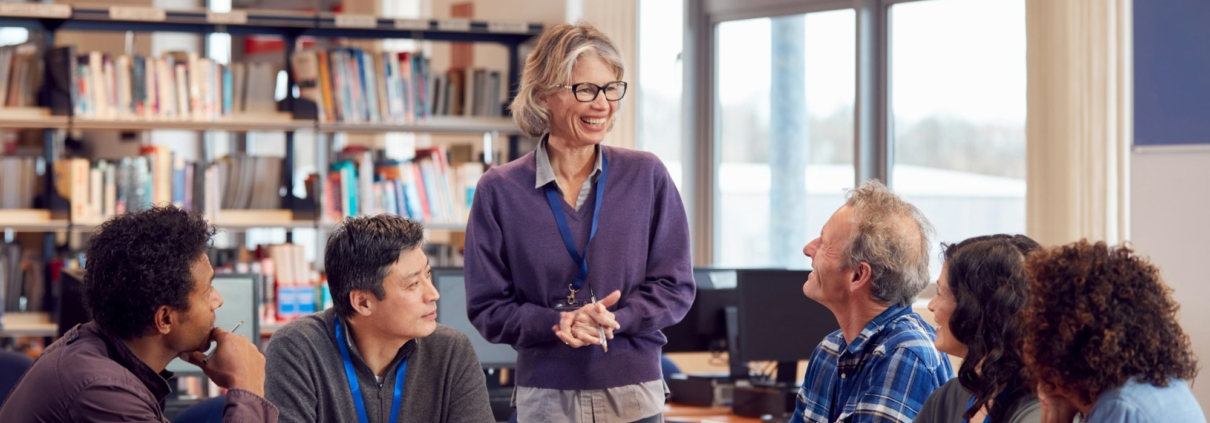 Teacher With Group Of Mature Adult Students In Class Sit Around Table And Work In College Library