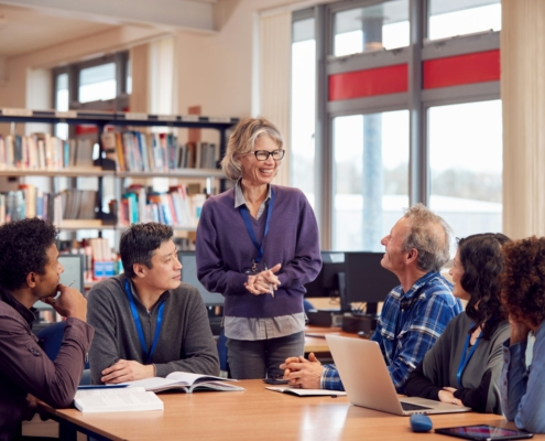 Teacher With Group Of Mature Adult Students In Class Sit Around Table And Work In College Library